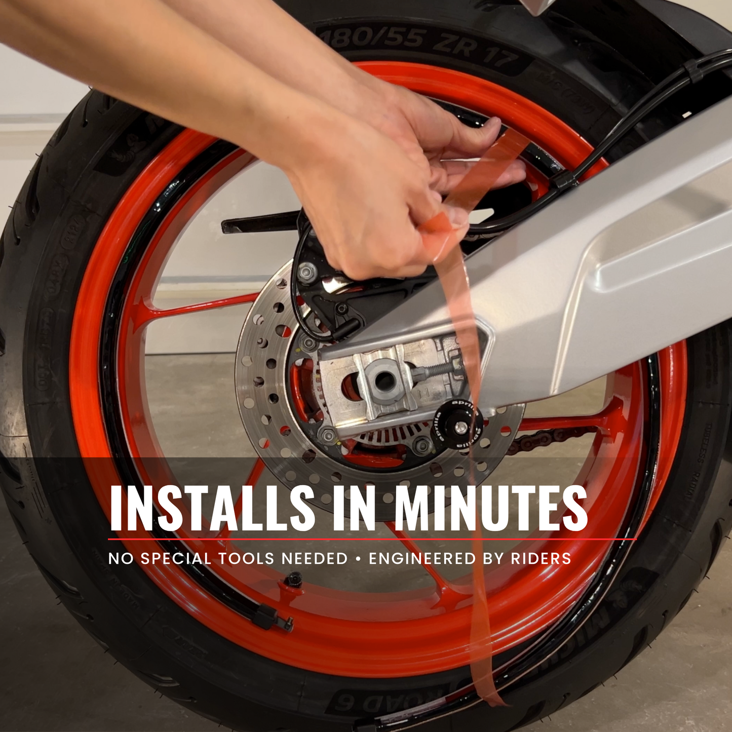 A close-up of a person's hands applying a peel and stick motorcycle wheel light to a motorcycle rim. The wheel features a bright orange rim, a silver fender, and a black brake disc.